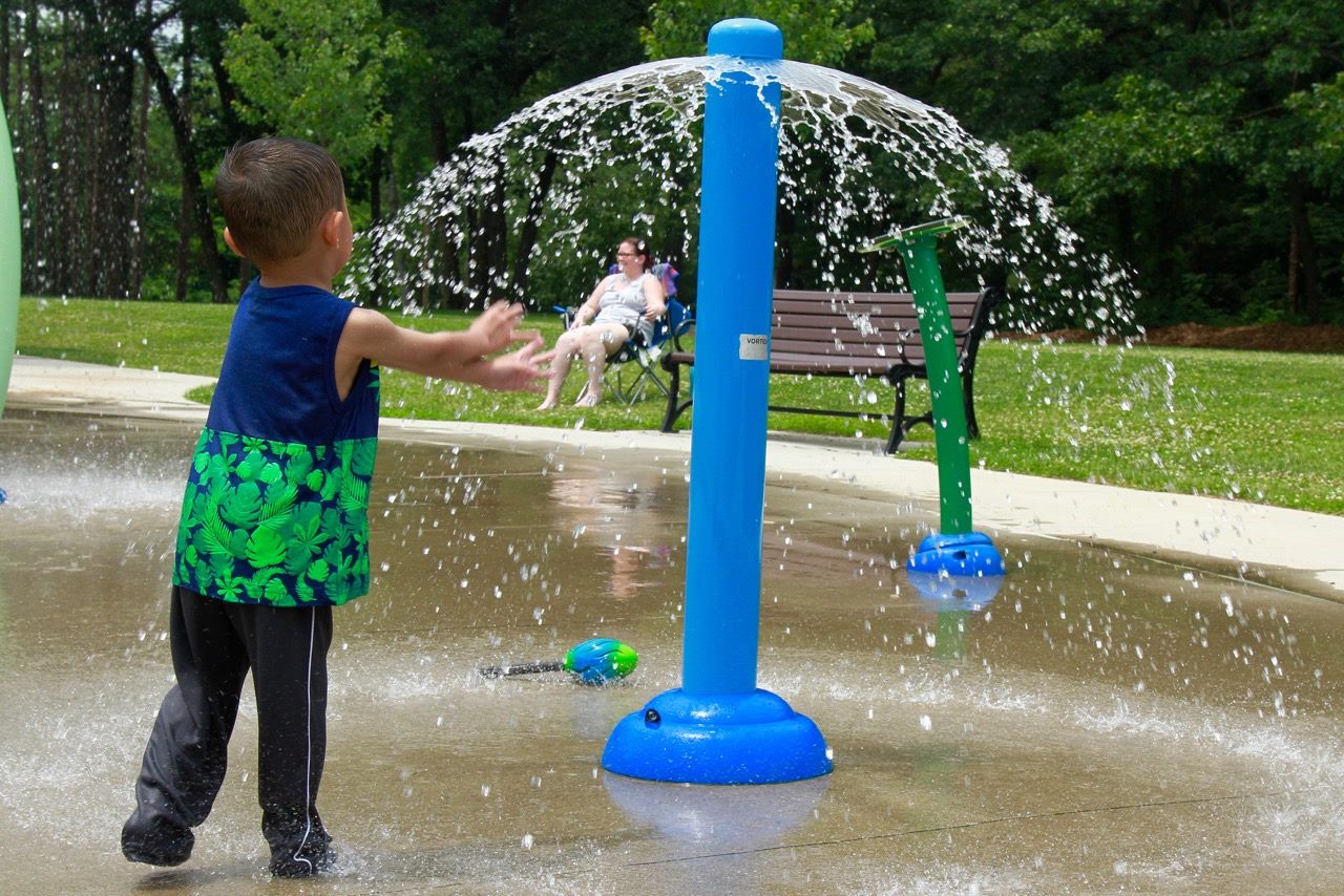 A small child runs toward a blue spray park bollard spraying water in a wide circle